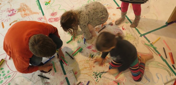 Various children's drawings on paper completely covering the floor for Drawing Disco at Southwark Park Galleries. Two children surrounded by pens are drawing with Alexa Lowe crouched next to them and one child standing.