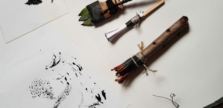 Five paintbrushes laid out one above the other made out of natural materials such as twigs and leaves on a white table. Left of the paintbrushes is a gestural painting in black using the paintbrushes on white paper.