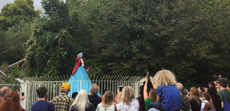 Marcia Farquar is performing on top of the old lido fountain outside Southwark Park Galleries, Lake Gallery. Marcia is wearing a blue bonnet, red open jacket and a blue skirt that flows over the three-tiered semi-circle fountain. In the foreground are lot’s of people watching.