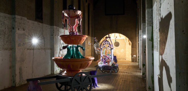 A concrete deconsecrated church is filled with a carnivalesque sculptures on wheels, brightly coloured and are spot lit, casting shadows onto the concrete walls.